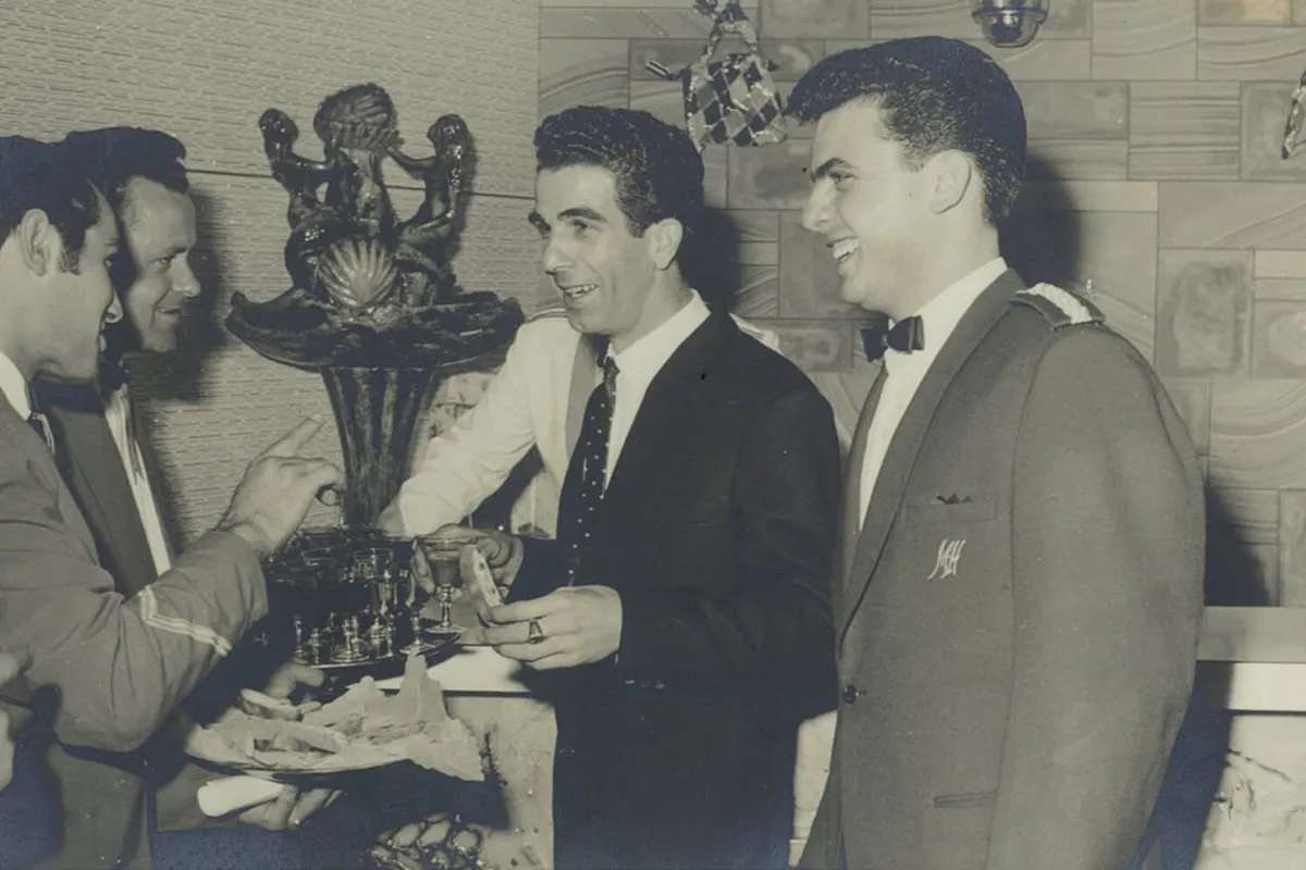 Black and white photograph of smiling men in formal attire gathered beside an ornate fountain, holding drinks and a plate of food.