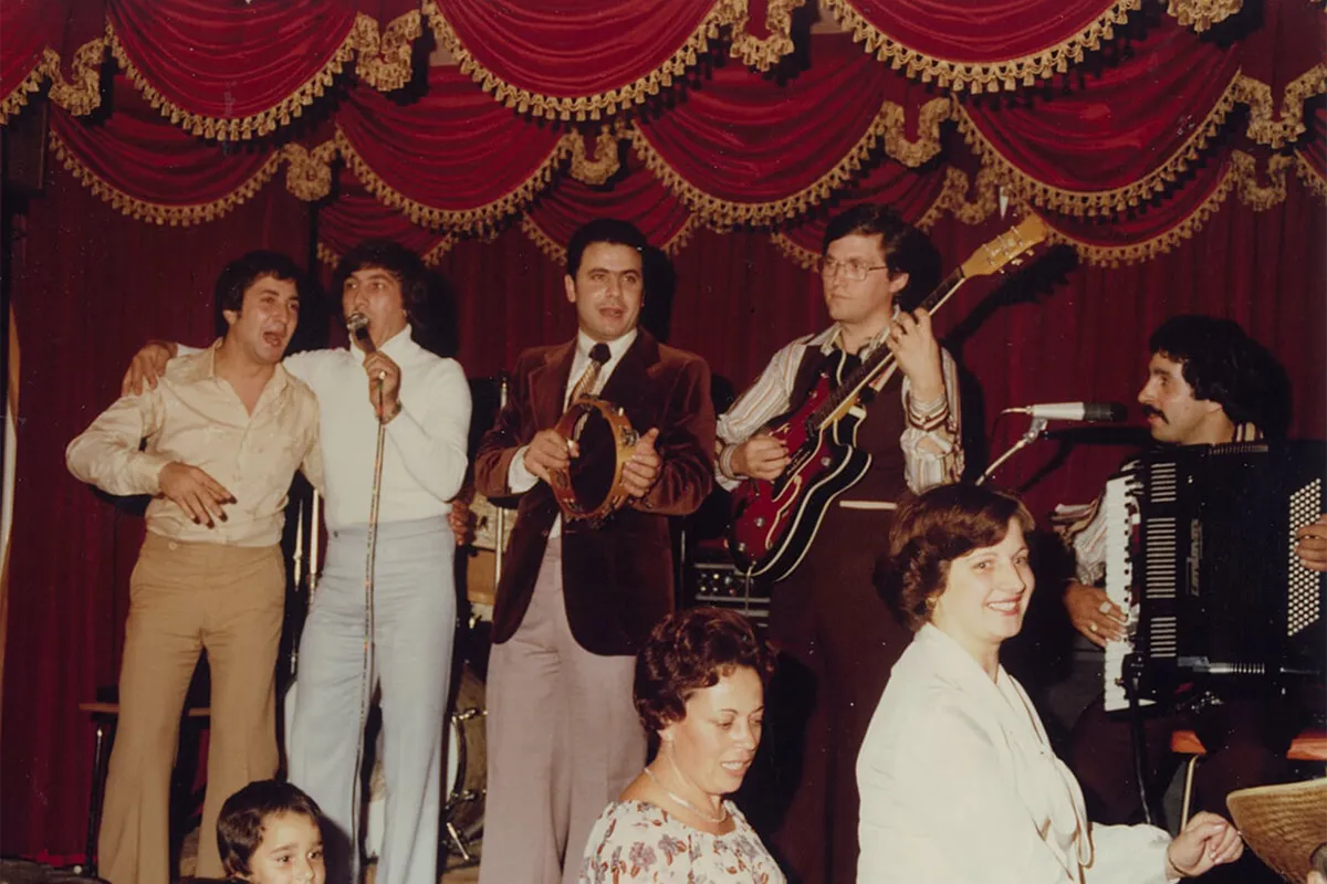 Singers and musicians performing with a tambourine, guitar and accordion in front of red velvet curtains.