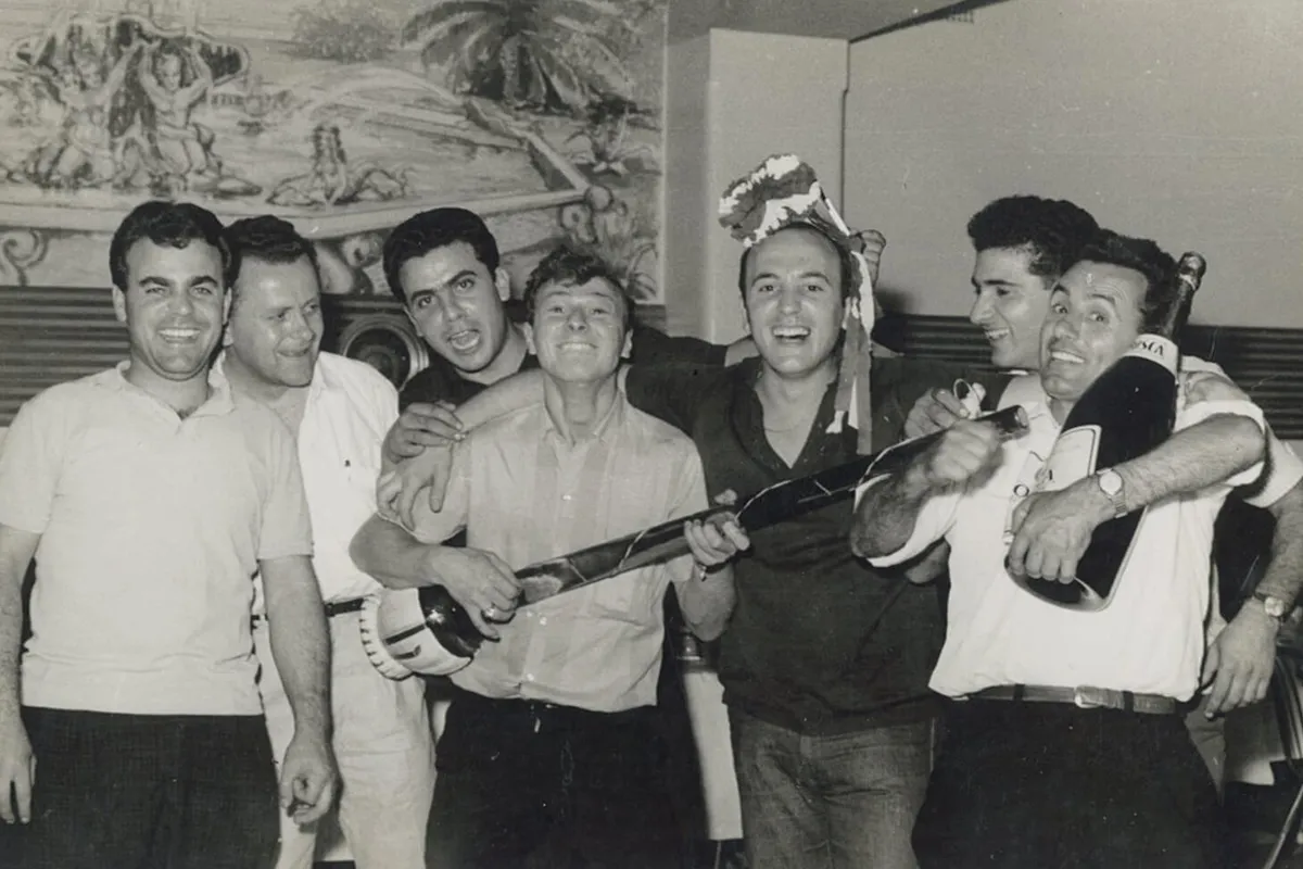 Black and white photograph of a group of men celebrating together, with one holding a guitar and another raising a tambourine.