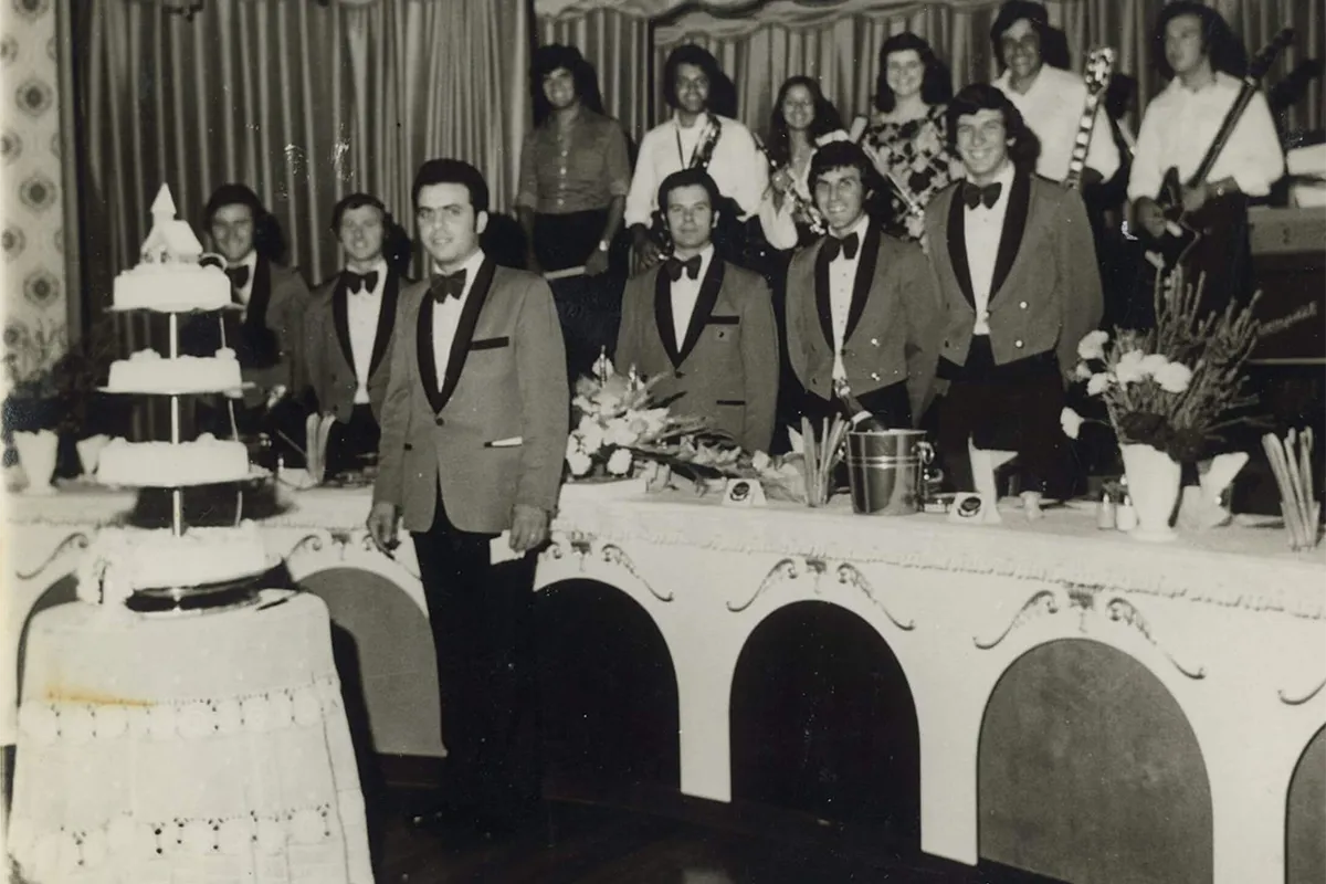 Black and white photograph of men in matching dinner jackets beside a tiered wedding cake, with musicians behind them.