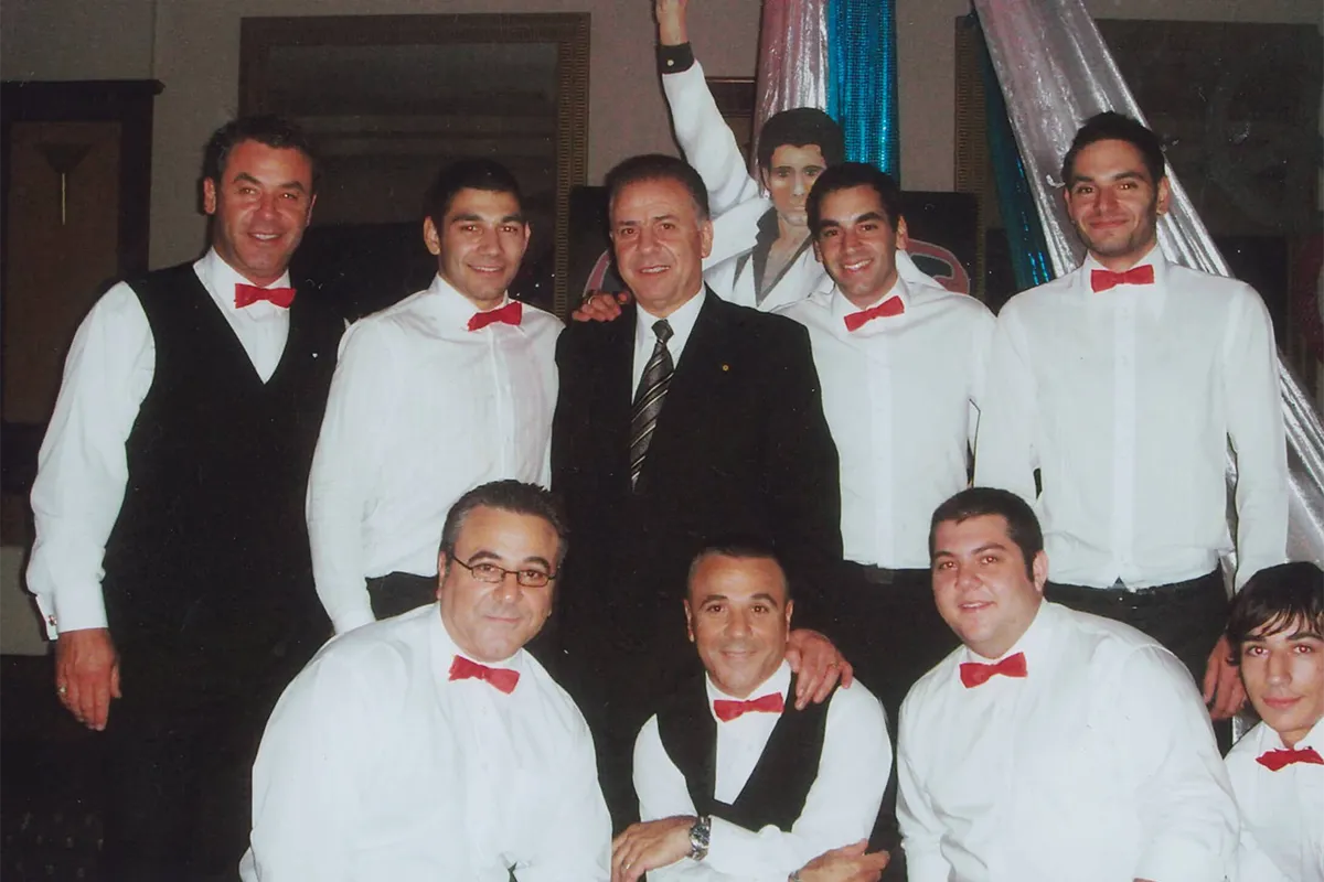Group portrait of men wearing white shirts and red bow ties gathered around a man in a dark suit.