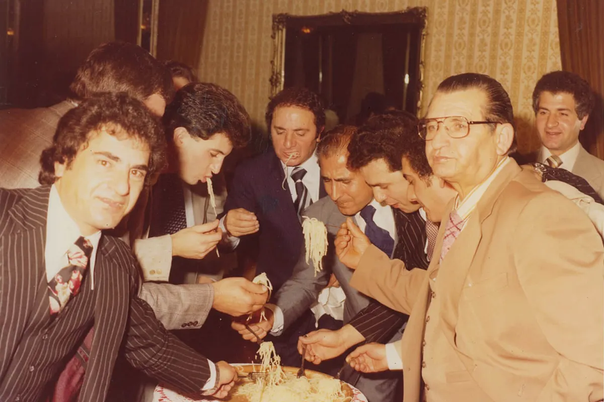 Men in suits gathered around a large platter, sharing spaghetti and smiling.