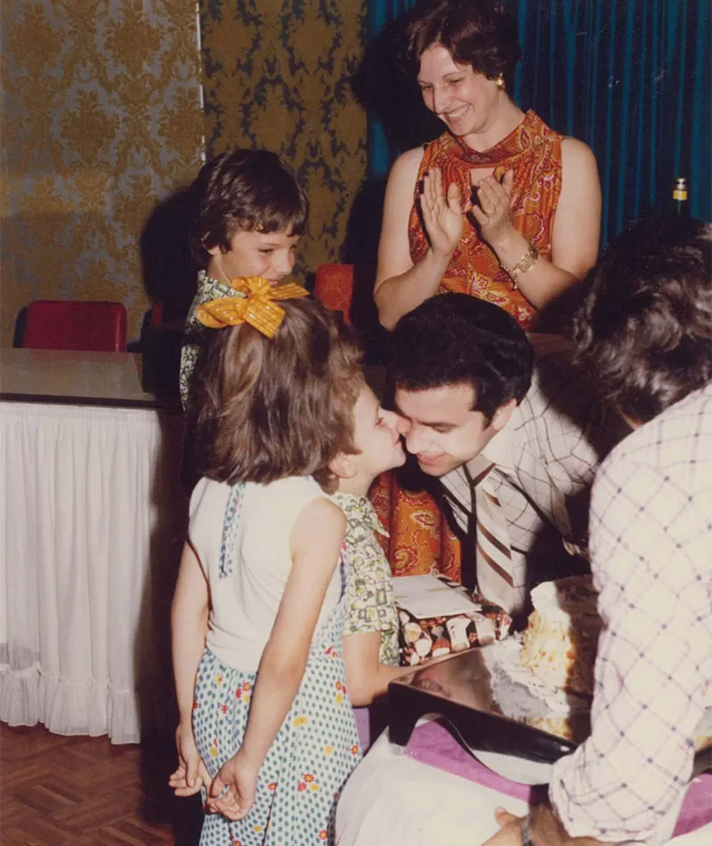 Filippo Navarra receiving a kiss from his granddaughter beside a celebration cake as family members look on.