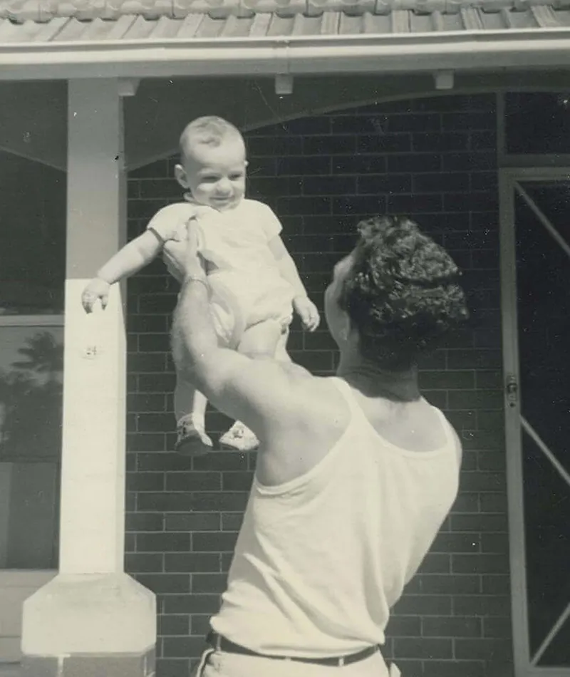 Black and white photograph of Filippo Navarra lifting his smiling young child outside a brick house.